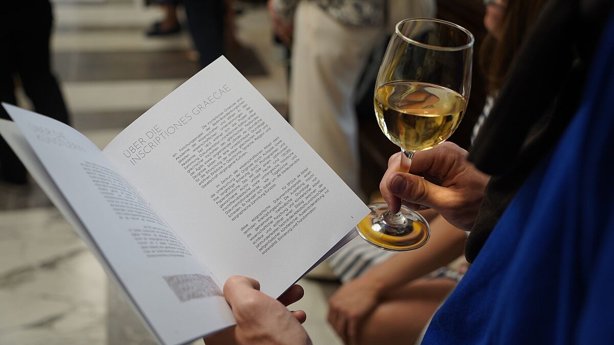 Nahaufnahme von einem Mann, der ein Booklet mit Text über die Inscriptiones Graecae hält. In der anderen Hand hält er ein Glas Weis