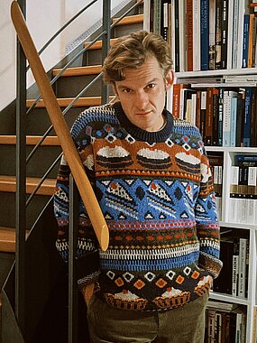 Andrei Zavadski poses infront of a bookshelf wearing a colorful jumper