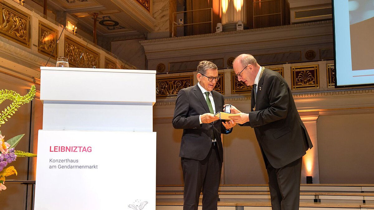 Auf der Bühne des prunkvollen Konzertsaals übergibt Christoph Markschies die Medaille an Reinhold R. Geilsdörfer. Neben ihnen steht ein Podium mit der Beschriftung "Leibniztag 2026"