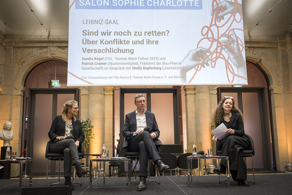 Drei Personen sitzen auf einem Podium auf der Präsentation der Schriftzug "Sind wir noch zu retten? Über Konflikte und ihre Versachlichung"