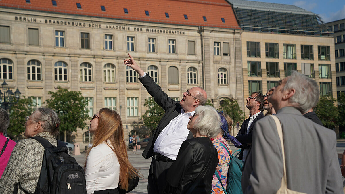 Akademiepräsident Christoph Markschies steht in einer Menschenmenge vor dem Gebäude der BBAW und zeigt aus dem Bild heraus auf den Dom..