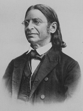 Foto von Abraham Geiger, ein Mann mit Nickelbrille und schulterlangem dunklen Haar, das er über den Koteletten zurückgekämmt hat. Er trägt einen schwarzen Anzug mit Weste.