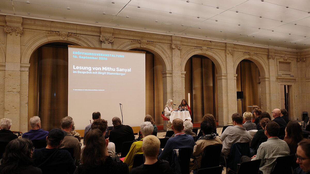 Auf der Bühne im Leibniz-Saal sitzen zwei Frauen vor vollem Publikum. Auf einem Screen sieht man den Schriftzug "Lesung von Mithu Sanyal im Gespräch mit Birgit Stammberger"