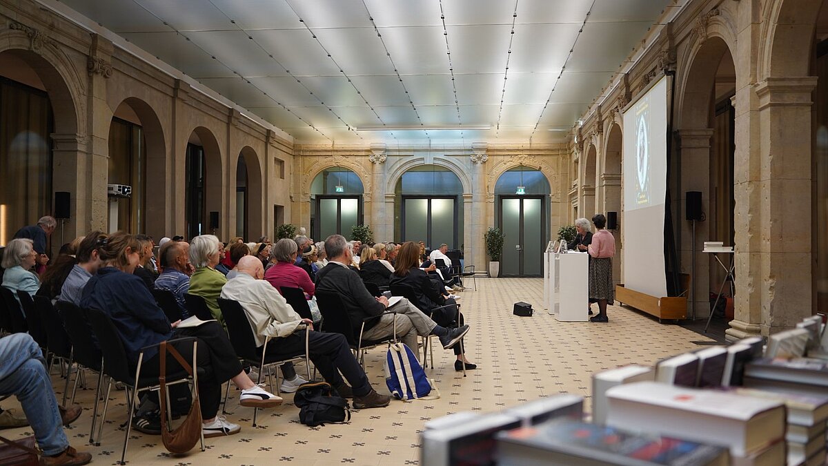 Der Leibniz-Saal ist voll. Menschen schauen zu, wie zwei Autor:innen ihre Bücher vorstellen. Im Vordergrund steht ein großer Stapel Bücher zum Verkauf.
