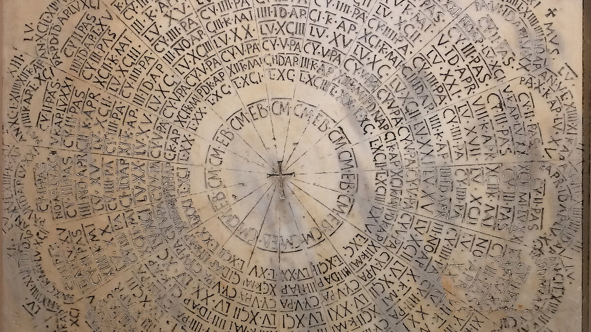 Marmorplatte mit konzentrischen Kreisen und Segmenten, die eine lateinische Inschrift tragen. In der Mitte befindet sich ein Kreuz, von dem Linien radial nach außen verlaufen. Die Inschriften sind in klaren, aufrechten lateinischen und gelegentlich griechischen Buchstaben ausgeführt und füllen die Segmente zwischen den Kreisen vollständig aus.