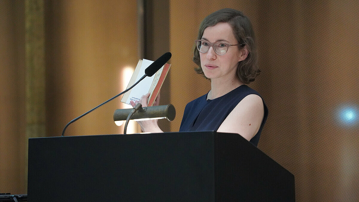 Marianne Seidig (eine Frau mit schulterlangen braunen Haaren) steht am Rednerpultund hält ein aufgeschlagenes Buch hoch.