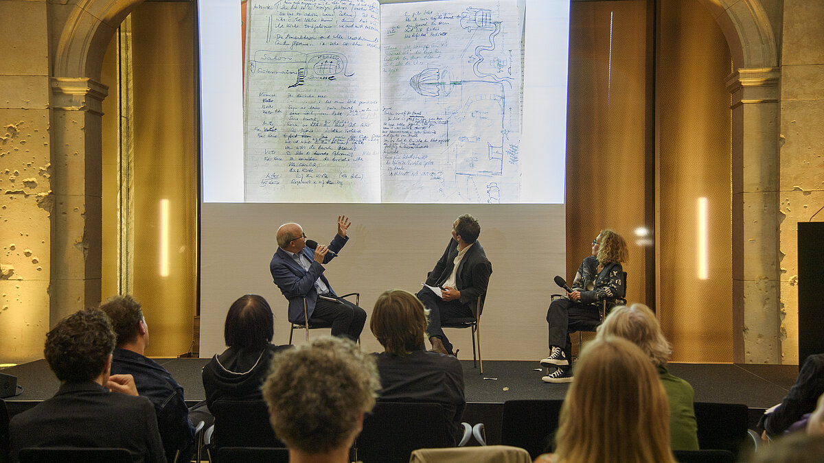 Drei Personen diskutieren auf einer Bühne im Leibniz-Saal. Eine Präsentation zeigt ein Foto von einem Notizheft mit Zeichnungen und schriftlichen Notizen