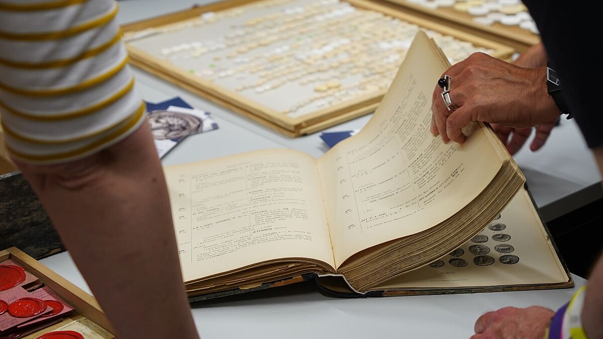 Zwei Personen blättern durch ein großes Buch über Münzen. Im Hintergrund sind viele Gipsabgüsse von Antiken Münzen zu sehen